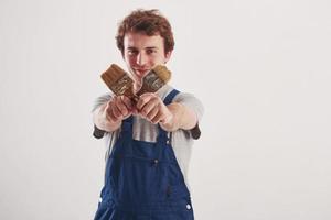 Crossed brushes in hands. Man in blue uniform stands against white background in the studio photo