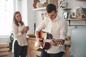 Girl is smiling. Young guitarist playing love song for his girlfriend in the kitchen photo