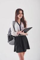 Homework goes well. Young female student in skirt and backpack with notepad in hands stands against white background photo