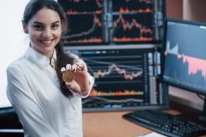 Nice daylight. Beautiful woman holding bitcoin in hands while standing in modern office with many monitors with graphs photo