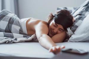 Smartphone addiction. Woman with blindfold mask for a sleep is lying on the bed at morning time photo
