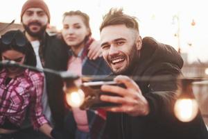 The wing is raised up guy's hair a little bit. Group of young cheerful friends having fun, hug each other and takes selfie on the roof with decorate light bulbs photo