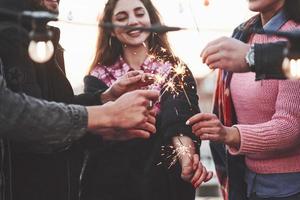 Smiling during the party. Playing with sparklers on the rooftop. Group of young beautiful friends photo