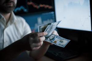 It's payday. Bearded man in white shirt counts money in the office with multiple computer screens in index charts photo