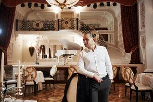 Satisfied look. Portrait of guy in official wear stands in beautiful restaurant in vintage style with piano on the stage photo