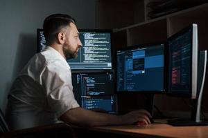 Adult experienced programmer. Bearded man in white shirt works in the office with multiple computer screens in index charts
