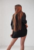 Rear view. Hot girl in black wear stands in the room against white wall photo