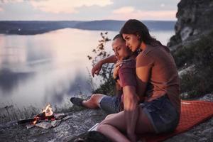 Hugging couple with backpack sitting near the fire on top of mountain enjoying view coast a river or lake. Traveling along mountains and coast, freedom and active lifestyle concept photo