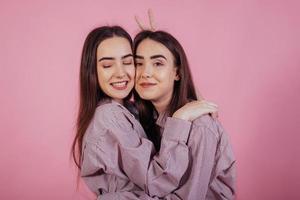 Girl from the left makes horn gesture behind other woman. Two sisters twins standing and posing in the studio with white background photo