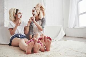 Laughing together. Pedicure and painted leg nails. Conception of skin care by using white mask and cucumbers on the face photo