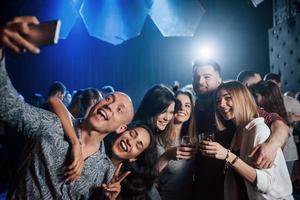 usando un teléfono inteligente de color plateado. amigos tomando selfie en una hermosa discoteca. con bebidas en las manos foto