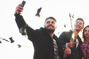 Attractive bearded man holds the phone. Group of young cheerful friends having fun while takes selfie on the roof with decorate light bulbs photo