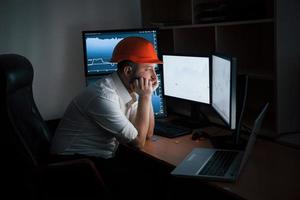 In the safety helmet. Bearded man in white shirt and red safety helmet works in the office with multiple computer screens in index charts photo