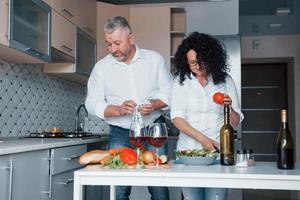 Clean and luxury interior. Man and his wife in white shirt preparing food on the kitchen using vegetables photo