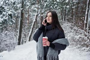 tener una conversación por teléfono. chica con ropa de abrigo con una taza de café da un paseo por el bosque de invierno foto