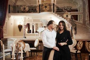 Man in white shirt, woman in black coat. Stands against stage with piano. Beautiful couple have romantic dinner in luxury restaurant at evening time photo