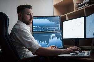 usando una computadora portátil de color plateado. hombre barbudo con camisa blanca trabaja en la oficina con múltiples pantallas de computadora en gráficos de índice foto