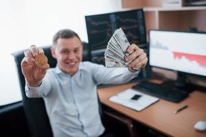 That's like success looks like. Businessman holding bitcoin and money in hands while sitting in modern office with many monitors with graphs photo
