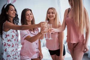 golpeemos los vasos. amigos celebrando la despedida de soltera en pijama en la sala blanca a la luz del día foto