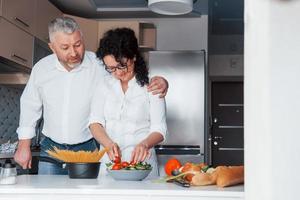 Love is like wine, getting better with every year. Man and his wife in white shirt preparing food on the kitchen using vegetables photo