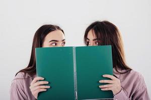 Looking at each other with wonder while holding notepad. Two sisters twins standing and posing in the studio with white background photo