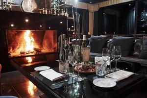 Wine glasses, bottles, white plates and delicious food. Interior of luxury restaurant in vintage aristocratic style with beautiful fireplace