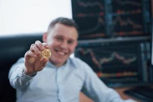 Happy young guy. Businessman holding bitcoin in hand while sitting in modern office with many monitors with graphs photo
