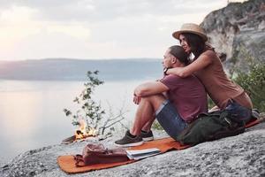 Hugging couple with backpack sitting near the fire on top of mountain enjoying view coast a river or lake. Traveling along mountains and coast, freedom and active lifestyle concept photo