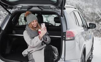 Taking a photo of nice snowy forest. Nice girl sitting at the rear part of modern car in the beautiful winter woods