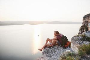 Hugging couple with backpack sitting on top of rock mountain enjoying view coast a river or lake. Traveling along mountains and coast, freedom and active lifestyle concept photo