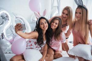 In the white room. Bachelorette party. Four girl in pink and white clothes sits at the sofa and take selfie photo