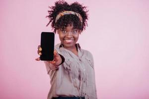 Smiling and feeling joy. Holding black phone in hand. Attractive afro american woman in casual clothes at pink background in the studio photo