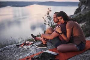 Hugging couple with backpack sitting near the fire on top of mountain enjoying view coast a river or lake. Traveling along mountains and coast, freedom and active lifestyle concept photo