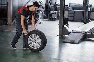 In red and black colored uniform. Mechanic holding a tire at the repair garage. Replacement of winter and summer tires photo