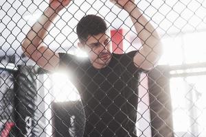 Sportive guy in black shirt stands behind the fence in the room with daylight behind photo