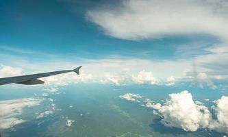 View from inside plane through plane window over blue sky, white clouds and green mountain. Commercial airline flight for summer travel. Plane wing above the city. Airplane flying above land. photo