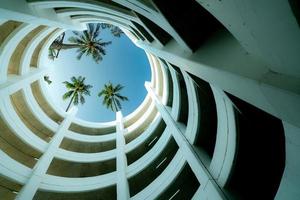 Bottom view multi-story car park building with coconut tree above building in summer. Multi-level parking garage. Indoor car parking lot. Architecture of spiral curve building. Sustainable building.