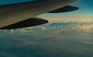 Wing of plane over mountain cover with white snow. Airplane flying on blue sky. Scenic view from airplane window. Commercial airline flight. Plane wing. Flight mechanics concept. International flight. photo