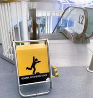 Beware uneven floor warning sign on yellow board in front of escalator in shopping mall. Beware uneven floor warning sign for safety at walkway before dawn way of escalator. Notice for safety. photo