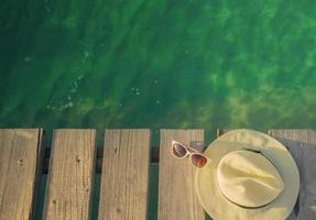 Top view of straw hat and sunglasses on wooden bridge over emerald green sea water. Summer vacation travel background. Summer vibes. Coconut tree shadow in sunglasses. photo