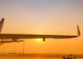 Closeup airplane wing with golden sunrise sky. Plane parked at airfield of airport. Morning flight. Summer travel concept. Pushback tractor support with airport staff at taxiways. Aviation business. photo