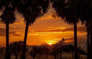 Beautiful orange sunrise behind the clouds. Silhouette old hut in forest near sugar palm tree in the morning. Golden sunrise sky. Country view. Sunrise shine with red and orange sky. Beauty in nature. photo