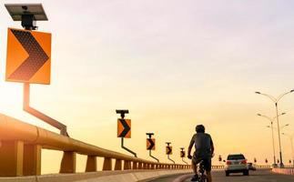 Back view of man wears helmet riding bicycle on the curve road behind cars. Safety driving and riding on the road. Curve road traffic sign with solar panel energy. Outdoor exercise. Healthy lifestyle. photo