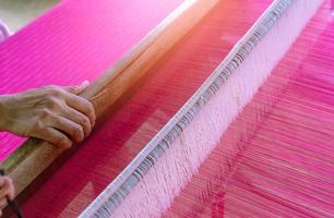 Woman working on weaving machine for weave handmade fabric. Textile weaving. Weaving using traditional hand weaving loom on the long cotton strands. Textile production in Thailand. Asian culture. photo