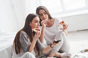 Girl licks her finger, it's so taste. Sisters eating pizza when watching TV while sits on the floor of beautiful bedroom at daytime photo