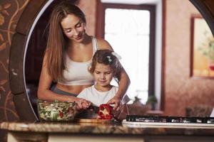 niño aprende rápido. una mujer muy joven parada en la cocina moderna cerca de la estufa de gas y le enseña a su hija a preparar comida foto