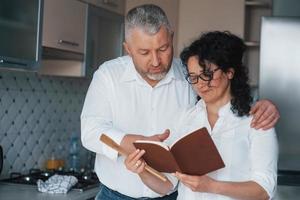 utilizando libro con recetas. hombre y su esposa con camisa blanca preparando comida en la cocina usando verduras foto