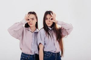 Gestures of rings on the eyes. Two sisters twins standing and posing in the studio with white background photo