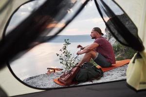 View from tent of traveller with backpack sitting on top of mountain enjoying view coast a river or lake. Traveling along mountains and coast, freedom and active lifestyle concept photo