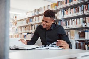 Smiling during the process. African american man sitting in the library and searching for some information in the books photo